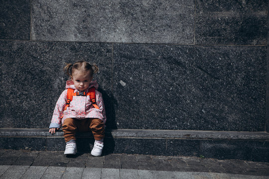 little pretty baby who got lost in the city, a girl in a pink raincoat, with a backpack and a stylish hairstyle sitting next to a large administrative building one by one