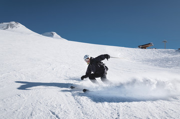A bearded mature aged male skier in a black ski suit descends along the snowy slope of a ski resort...