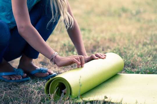 Cropped image of girl rolling yoga mat. Close-up of attractive young woman folding green yoga or fitness mat after working out in a glade in the woods or a city Park . Healthy life