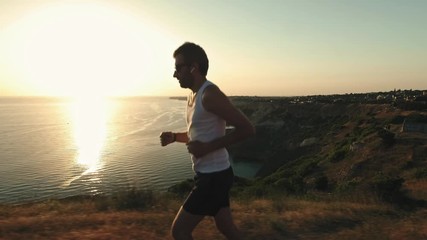 Athletic Handsome Man is Doing Sports with Phone and Headphones, Running along Sea Coast at Sunset while Jogging on Beach in Summer. Uses wireless headphones
