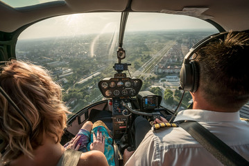 Portrait of beautiful blonde women and pilot enjoying helicopter flight. She is amazed by cityscape. - obrazy, fototapety, plakaty
