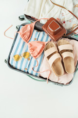 Flat lay of luggage with summer clothes and accessories: bikini, slippers, retro camera, sunglasses, dress on white background. Top view fashion travel composition.