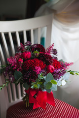 A beautiful wedding bouquet in red-burgundy tones stands on a chair. Hands. Red roses and peonies. Wedding day. Bokeh. Bride. Bride's bouquet. Wedding flowers. The bouquet is decorated with a brooch. 