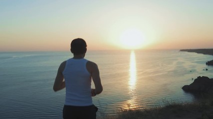 Athletic Handsome Man is Doing Sports with Phone and Headphones, Running along Sea Coast at Sunset while Jogging on Beach in Summer. Uses wireless headphones