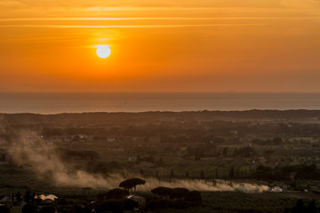 The sun sets on the coast of Tuscany, Italy, while the smoke of a fire lit in the hinterland rises