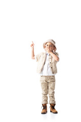 full length view of shocked explorer boy in hat and glasses showing idea sign and covering mouth with hand on white