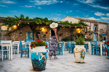 Young romantic woman walks near the colorful outdoor cafe  in the beautiful sicilian coastal village Marzamemi in Sicily, south Italy - obrazy, fototapety, plakaty