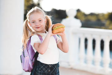 Cute child girl 5-6 year old holding hamburger outdoors. Wearing school bag and uniform close up. Back to school. 1 September.