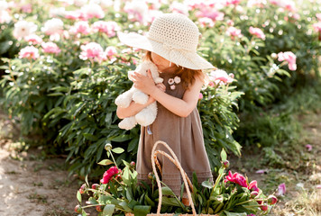 pretty girl child in a straw hat plays with a soft toy a lamb in the garden of blooming peonies