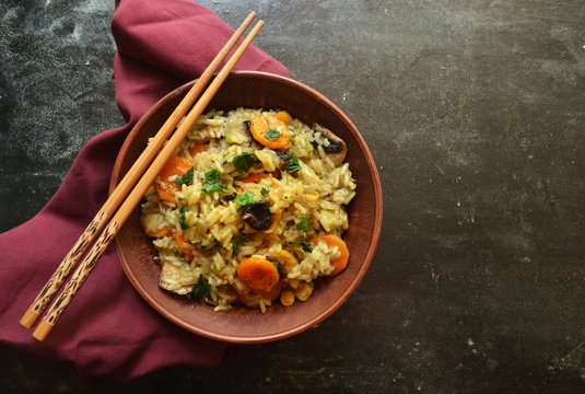 Photo of food. Appetizing rice with vegetables in a brown bowl. Rice with mushrooms, herbs and carrots. Food on a black background isolated with burgundy textile.