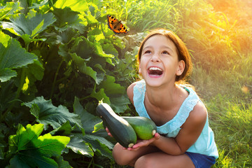 Happy laughing girl holding fresh zucchini outdoors in garden looking on flying butterfly