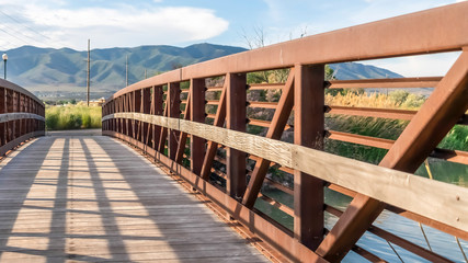 Fototapeta na wymiar Panorama frame Wooden bridge with metal lattice guardrail over a lake with view of mountain