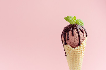 Chocolate brown ice cream scoop in waffle cone with chocolate sauce, mint leaf on pink background,...