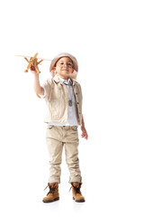 full length view of smiling explorer boy in glasses and hat holding wooden toy plane isolated on white