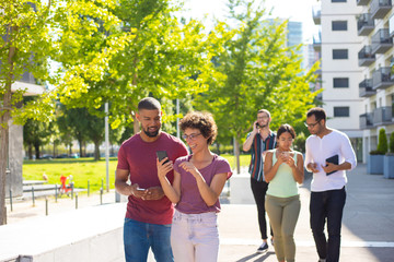 Obraz premium Happy young people using mobile phones while walking through residential area. Men and women talking on cell, using smartphones and showing screen to each other. Wi-Fi outdoors concept