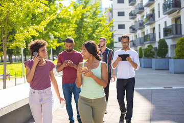 Group of young people using digital devices while walking outside. Young men and women talking on cellphones and using smartphones and tablet. Mobile technology concept
