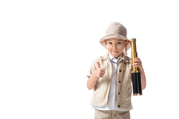 front view of smiling explorer boy in glasses and hat holding spyglass and showing thumb up isolated on white