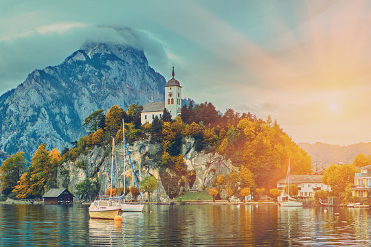 Beautiful scenic sunset over Austrian alps lake. Boats, yachts in the sunlight infront of church on the rock with clouds over Traunstein mountain at the alps lake near Hallstatt Salzkammergut Austria