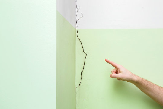 Man's Hand Finger Pointing To Cracked Corner Wall In House. Building Problems And Solutions Concept. Close Up.