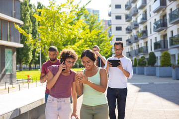 Two women walking outside together, talking on cell and staring at phone screen. Group of young men using gadgets behind them. Digital technology concept