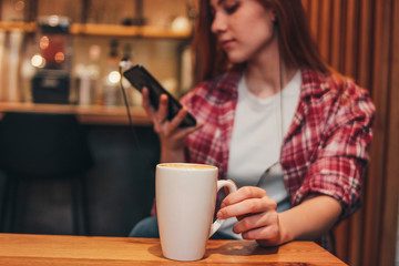 Cup of coffee cappuccino in female hands at cafe. Young woman in casual using mobile on background