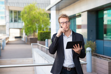 Serious businessman using tablet and calling on phone. Young business man standing outside, holding mobile computer and talking on cell. Wireless connection concept