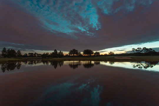A sunrise over a golf course lake, with clouds in the sky providing a mix of orange and pink light behind the distant trees and reflection on the water.