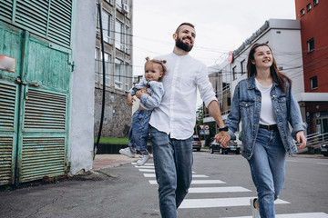 Obraz premium concept of a young happy family with one child: a stylish bearded father, a beautiful young wife and their smiling 2-year-old daughter walking in the city on their hands in the city