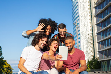 Happy friends using video call app on tablet. Mix raced team of people sitting outside, smiling, shouting, laughing, waving hello to screen. Video talk concept