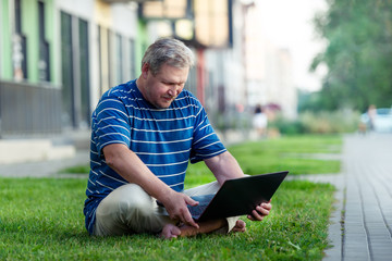 Happy man looking for online content on laptop sitting on lawn on summer day in city