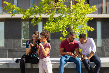 People sitting on parapet outside and using smartphones. Men and women excited with their phones and discussing content. Phone using concept