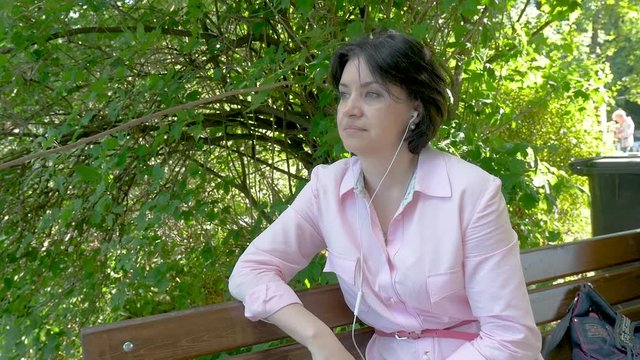 A middle-aged brunette woman in a pink blouse with a clock on her hand uses white headphones to listen to music. She is sitting on a bench near the pond on a summer sunny day. close-up