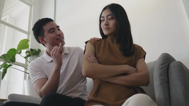 Young Asian man trying to reconcile to girlfriend while sitting on the sofa at home.