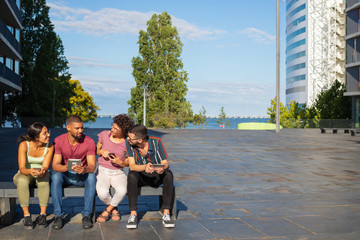 Happy young men and women using mobile gadgets and discussing content. People sitting on bench outside and using smartphones and tablets. Communication concept