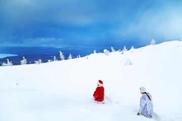 Authentic Santa Claus and a girl in winter clothes are walking on a snowy mountain.