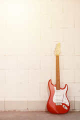 Fototapeta na wymiar Red electric guitar stands on the right against white brick wall, yellow sunbeam effect