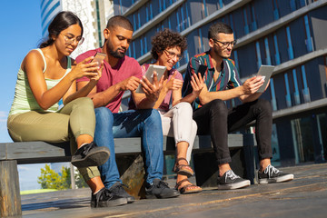 Obraz premium Group portrait of young people focused on their digital gadgets. Men and women sitting on bench outside, using mobile app or consulting internet. Communication concept