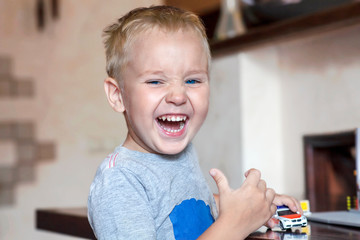 Little caucasian baby boy with blond hair and bright blue eyes laughs holding the car toy and showing thump up. Strong emotions. Indoors, copy space.
