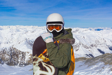 Snowboarder man with orange ski glasses in white helmet posing on top in Alps mountains. On the background of mountains.