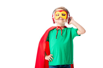 front view of smiling preschooler kid in mask and red hero cloak listening music in headphones and looking at camera isolated on white