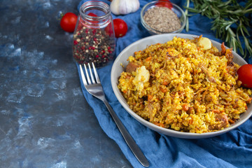 A large plate filled with Uzbek pilaf, next to cherry tomatoes, a dark cloth, fresh pepper in a manually knitted stand. In a transparent dish, spices - zeros and a mixture of peppers. 