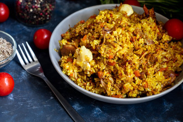 A large plate filled with Uzbek pilaf, next to cherry tomatoes, a dark cloth, fresh pepper in a manually knitted stand. In a transparent dish, spices - zeros and a mixture of peppers. 