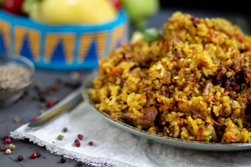 A large plate filled with Uzbek pilaf, next to cherry tomatoes, a dark cloth, fresh pepper in a manually knitted stand. In a transparent dish, spices - zeros and a mixture of peppers. 