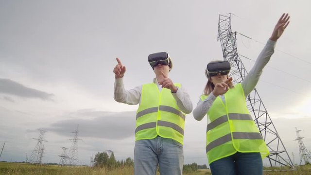 A man and a woman engineer in VR glasses control the power distribution of electric networks and the delivery of electricity against the background of electric towers with high-voltage cables.