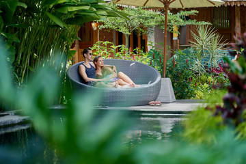 Candid shot of young caucasian couple lounging in modern chair near luxurious pool of tropical hotel and spa in Bali