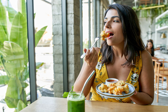 Candid lifestyle portrait of fashionable healthy ethnic businesswoman eating an organic vegetarian cauliflower dish for vegans and drinking green smoothie