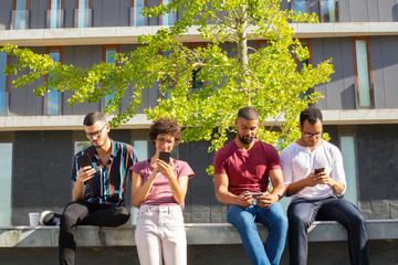 Four friends totally excited with their mobile phones. Men and woman standing outside, sitting on parapet and using smartphones. Internet addicted concept