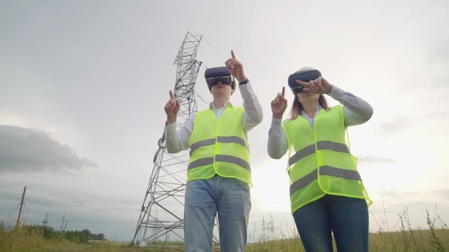 A man and a woman engineer in VR glasses control the power distribution of electric networks and the delivery of electricity against the background of electric towers with high-voltage cables.