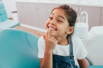 Rolgordijnen Tandarts Kleine patiënt van gemengd ras die haar perfecte brede glimlach laat zien terwijl ze op de stoel van de tandarts zit