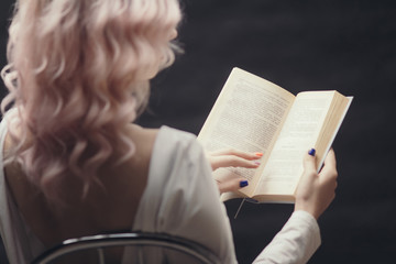 blonde girl holding open book sitting on a chair, back ofyoung woman reading a novel, concept of hobby, relaxation, leisure on black background